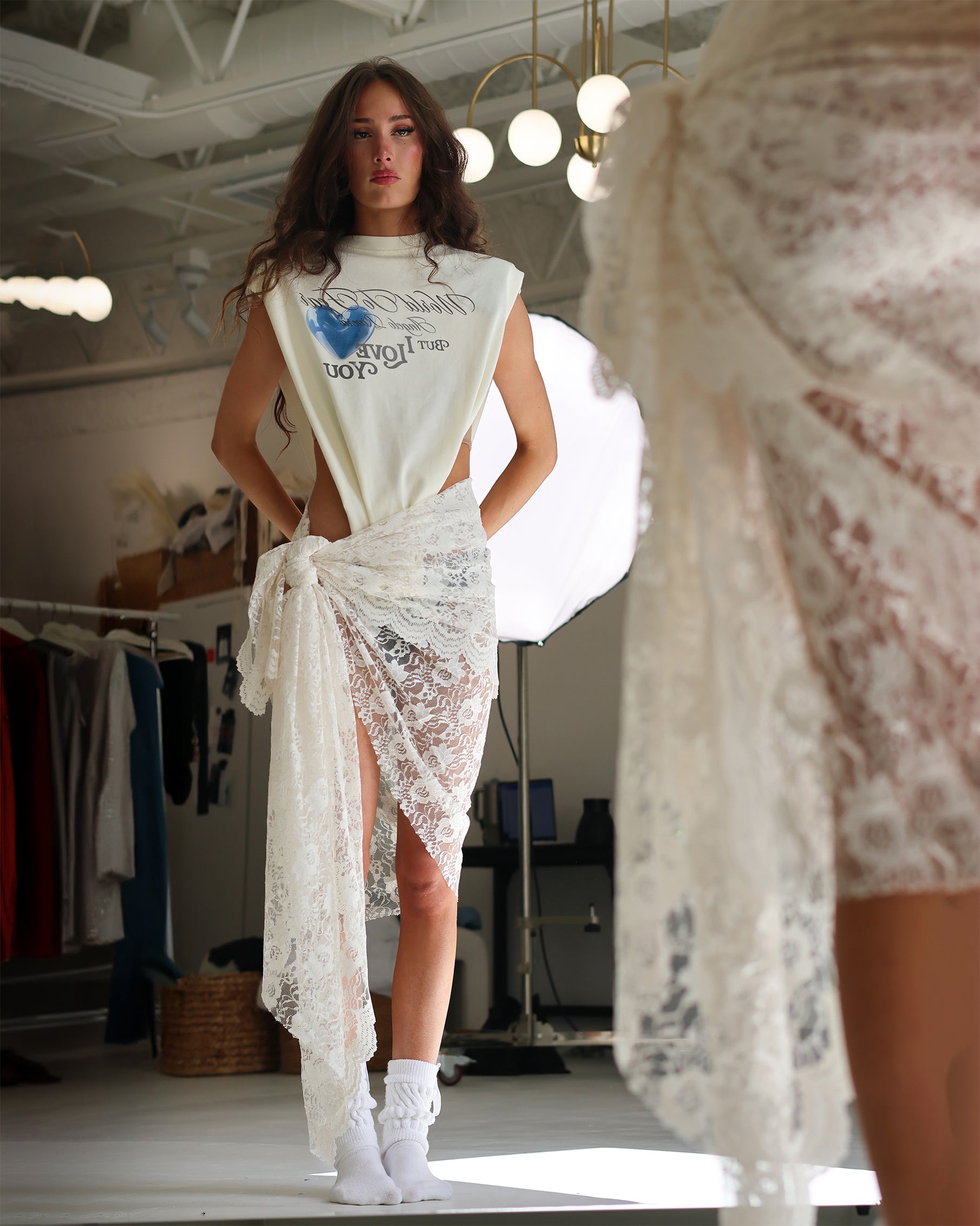 Model wearing a white dress with lace detailing in a room with clothing racks.